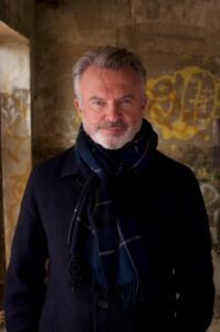 Portrait of a middle-aged man with gray hair and beard, wearing a navy scarf and dark coat, standing under a graffiti-covered wall.